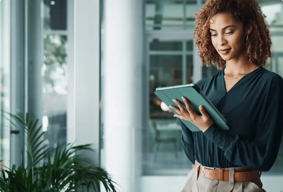 Woman using tablet indoors near plants and windows.