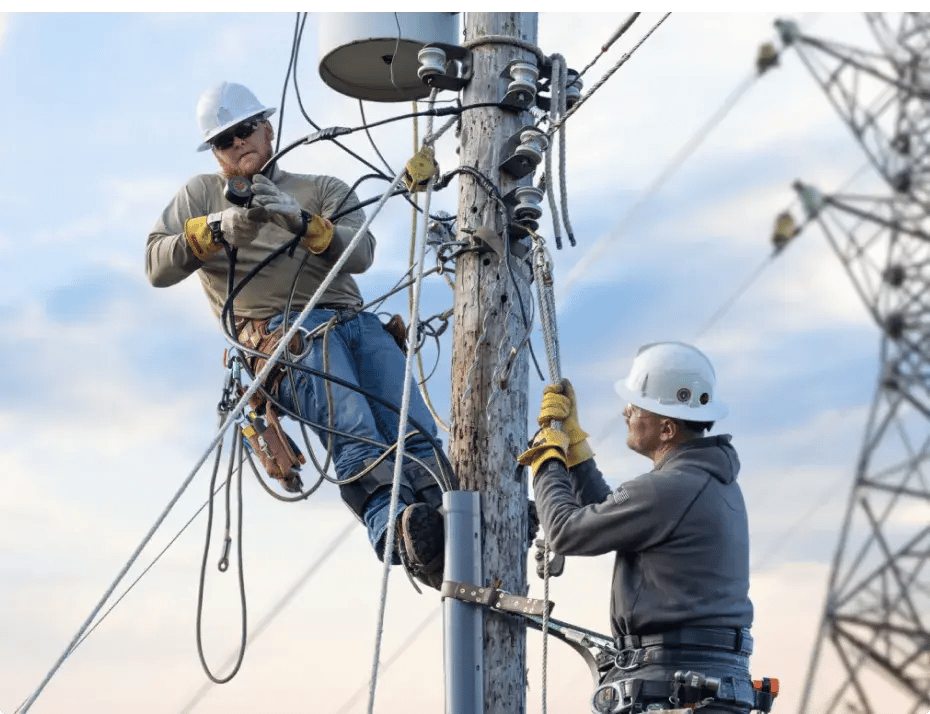 Electricians work atop a bucket lift at dusk, trimming branches near power lines.