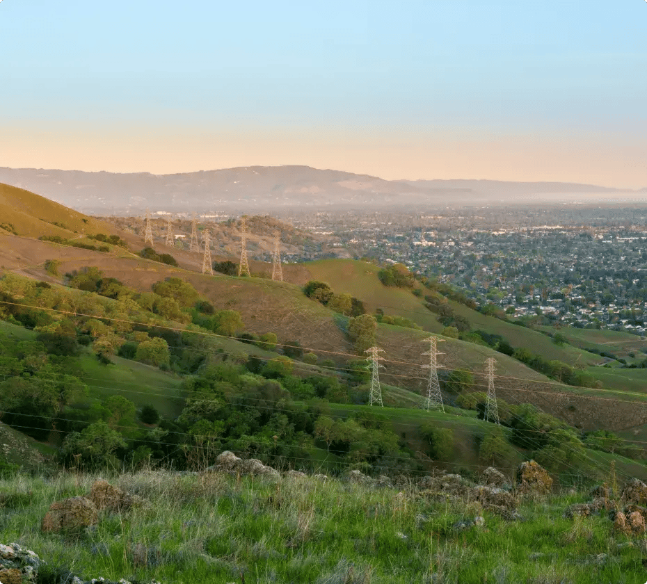 Scenic view of rolling green hills with power lines and transmission towers stretching across them, overlooking a hazy urban cityscape in the valley below at sunset. Distant mountains under a clear sky with warm orange hues.