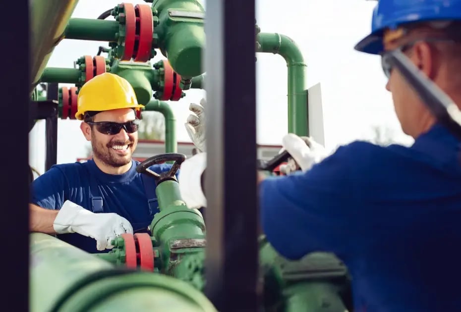 Two workers in blue uniforms and hard hats operate green valve systems with red handles.