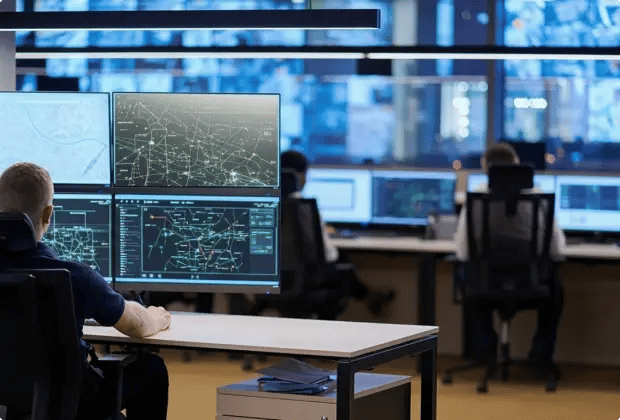 People working in a control room with multiple large monitors showing maps, network diagrams, and system data.