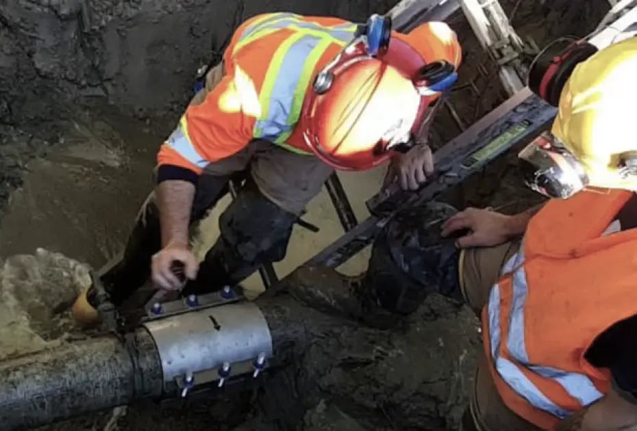 Workers in safety gear repair a pipe in a muddy trench using tools.