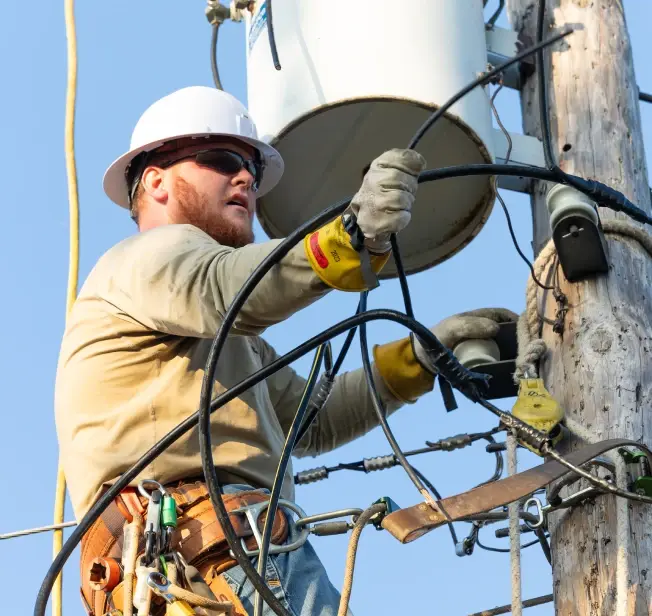 A utility lineworker wearing a hard hat, safety glasses, gloves, and a tool belt works on overhead electrical equipment from a bucket truck, handling cables and components on a wooden utility pole.