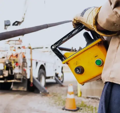 Worker with control box near utility truck and traffic cone.