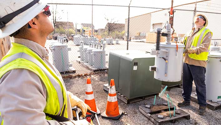 Two electric utility workers operating a crane and lifting a capacitor off the ground