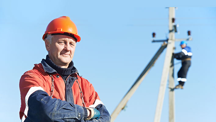 A utility worker posing with his arms crossed while another works on a telephone pole in the background