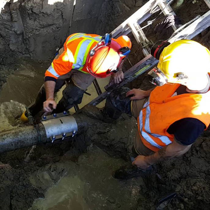 Two utility workers standing in a dugout hole, leaning down to inspect a pipeline