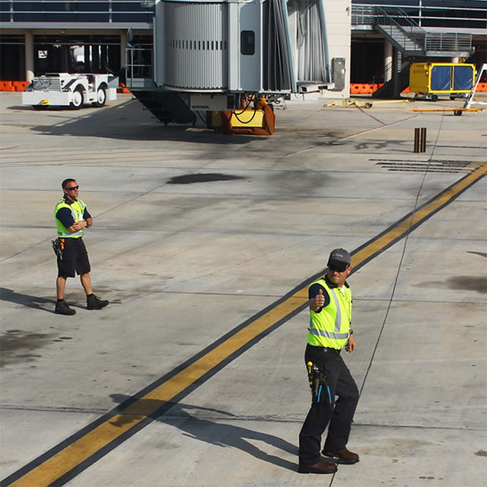 Two utility workers near a jetway guiding an offscreen airplane towards the bridge