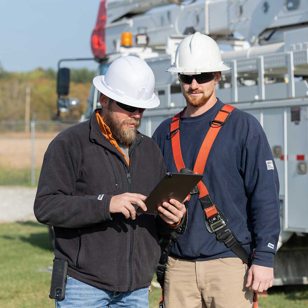 Two utility workers standing next to each other, one holding a tablet and the other looking down at it