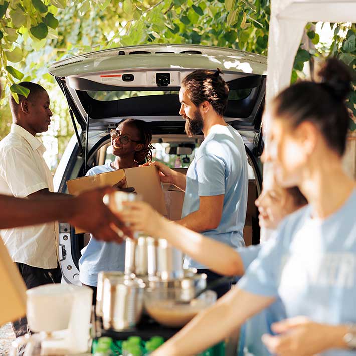 A group of people handing out food from the back of a car, under a tree